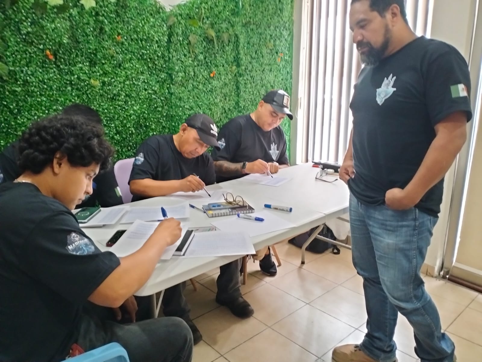 Alumnos con playera negra tomando notas en el aula durante la teoría, con el instructor de pie a su lado
